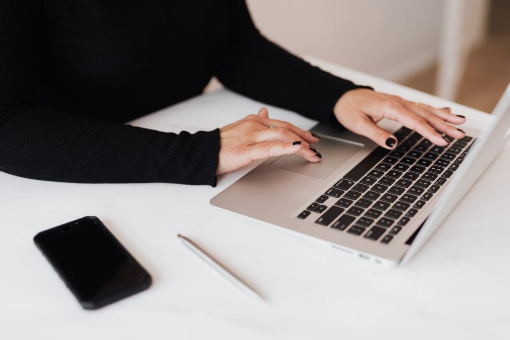 A woman typing on a laptop with a cell phone.