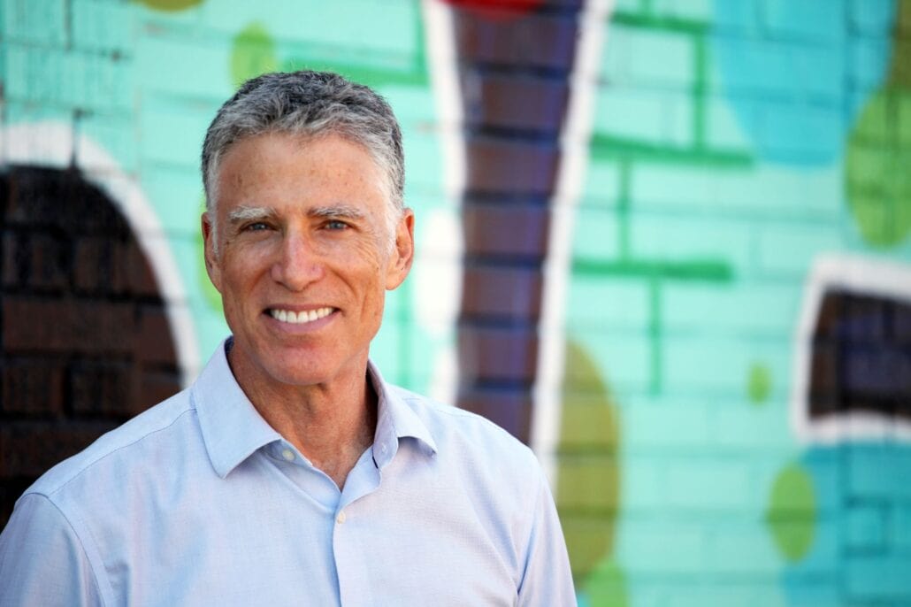 A man with gray hair wearing a light blue shirt smiles in front of a colorful mural on a brick wall.