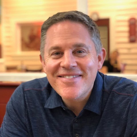 Chris Gargano A man with short brown hair and a blue collared shirt smiles while sitting indoors at a restaurant with warm lighting.