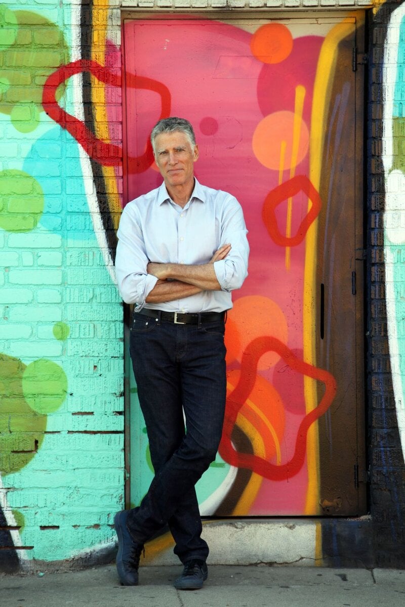 JT-7451-1000 A man with gray hair, wearing a light blue shirt and dark jeans, stands with arms crossed in front of a colorful graffiti-covered door.