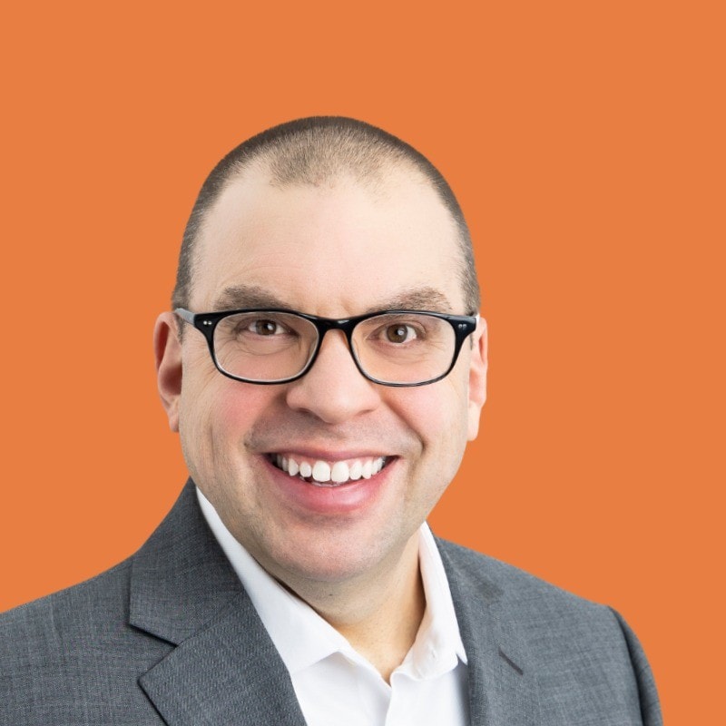 Jim Lombardi A man wearing glasses, a gray suit, and a white shirt smiles at the camera against an orange background.