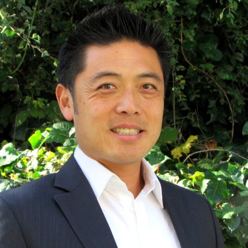 John Chen A man in a dark suit and white shirt stands outdoors in front of green leafy bushes, smiling at the camera.
