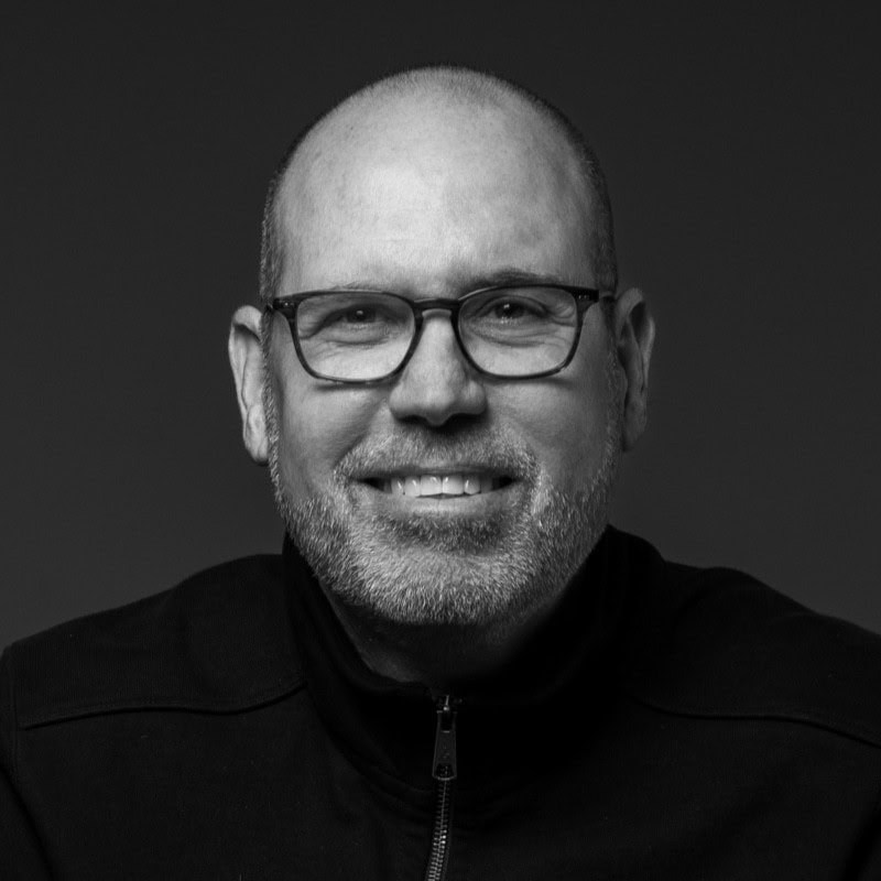 Mike Gollom Black and white portrait of a middle-aged man with glasses, a trimmed beard, and a bald head, smiling and wearing a dark zip-up jacket.